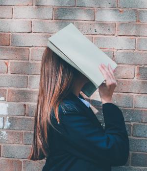 Pupil with book over face in front of brick wall